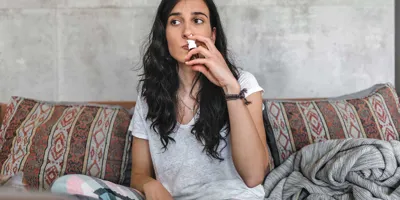 Koivun siitepölyallergiasta kärsivä nuori nainen istuu sohvalla pidellen nenäsumutetta nenäänsä vasten