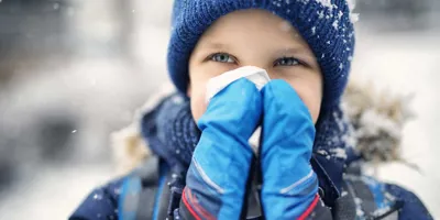 Photo d’un enfant qui se mouche. Il est dehors sous la neige et porte un bonnet et des moufles.